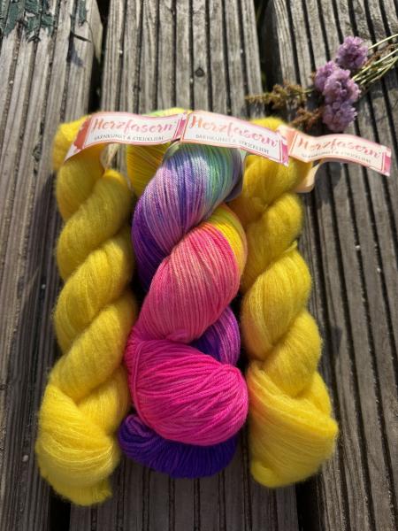 Handgefärbtes Petit Provence Garnset von Herzfasern mit einem bunten Strang „Yolli“ auf Herzfasern No. 11 sowie zwei fluffigen Strängen Herzfasern No. 93 in leuchtendem Zitronengelb auf einer Holzbank im Sonnenlicht fotografiert.
